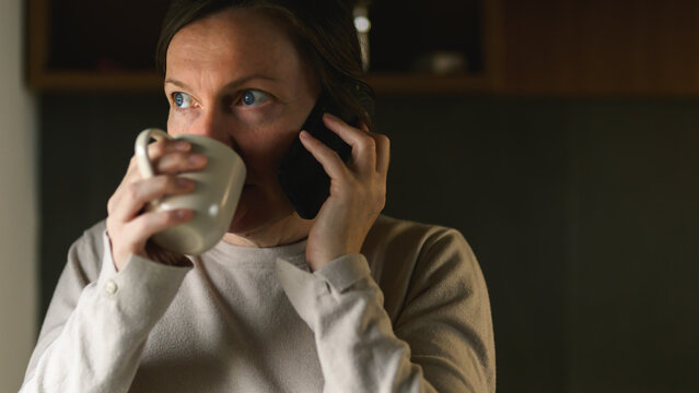 Woman drinking coffee and talking on phone in kitchen during quiet morning moment