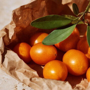 Fresh kumquats with green leaves in crumpled kraft paper bag, warm sunlight, organic citrus market harvest concept