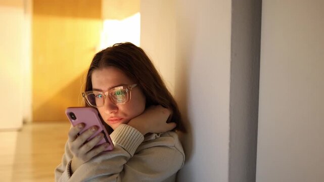 Woman leaning on wall at home scrolling smartphone instead of doing household tasks looking focused but detached from reality illustrating digital distraction and passive content consumption