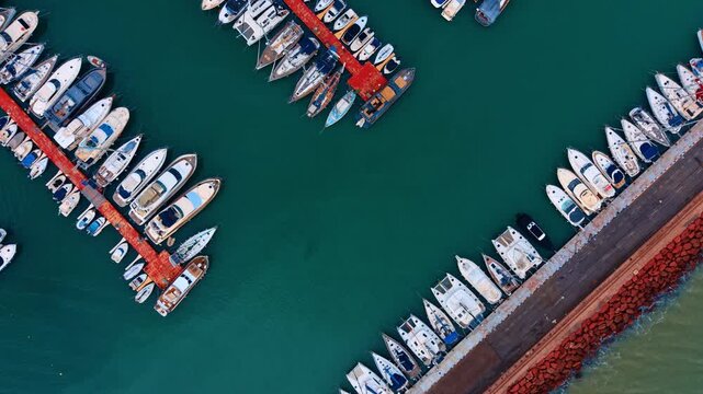 Aerial top view of white boats moored at marina pier. Top down drone shot of yachts and motorboats docked at a red pier in turquoise sea water.
