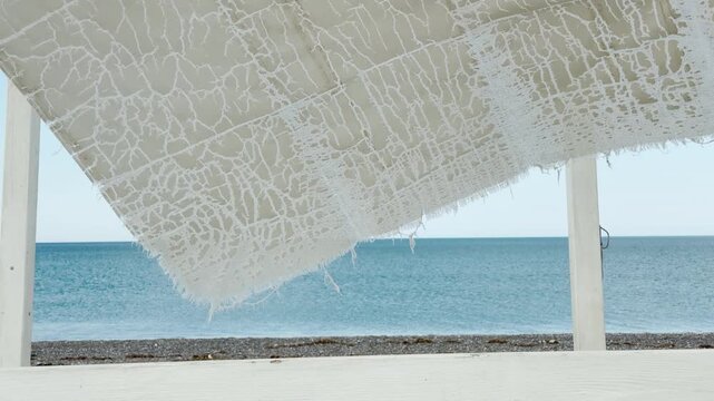 A shredded fabric canopy stretches above the empty shore facing the calm ocean. The worn texture against the water creates a feeling of peaceful solitude and nostalgia.