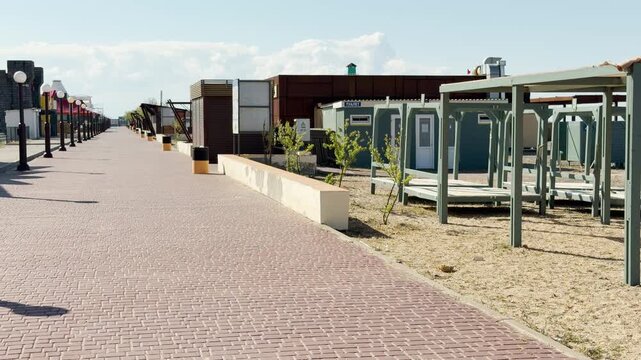 A quiet stone walkway stretches past wooden structures on a sandy coast. The empty path gives a feeling of peaceful solitude and anticipation before the arrival of summer visitors.