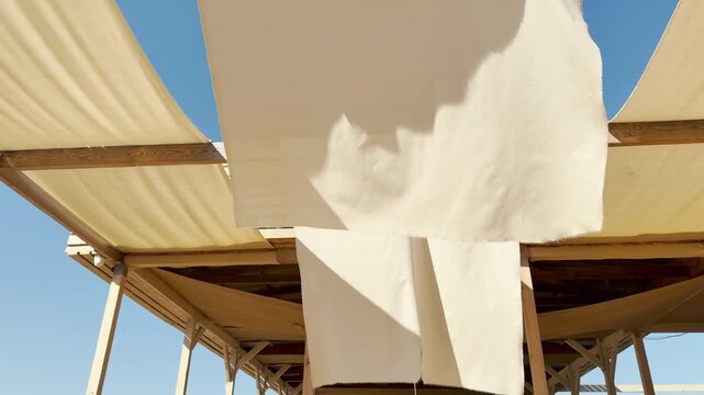 Soft white fabric panels flutter gently on a wooden frame against the sky. The warm sunlight and moving shadows create a peaceful, relaxed mood perfect for a coastal holiday.