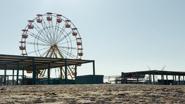 A tall Ferris wheel stands over a quiet beachside amusement park under a clear sky. The empty seaside setting creates a feeling of peaceful solitude and nostalgic calm.