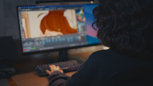 Rear view of middle-aged female artist with curly hair in sharp focus, blurred background showing hand typing on keyboard and monitor with cartoon character illustration. Cozy loft, warm evening light