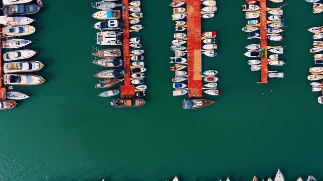 Directly above top down view of yachts in a marina. Symmetrical top down view of white boats docked at red piers in emerald green water.