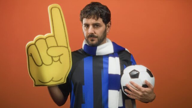 Man with beard holding soccer ball and foam finger, hand gripping ball and foam finger raised, wearing blue team scarf in studio; team pride.