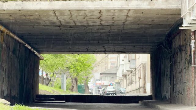 Heavy raindrops fall through a dark, weathered concrete tunnel. The gloomy passage opens up to a cheerful urban street filled with vibrant green trees and passing cars.