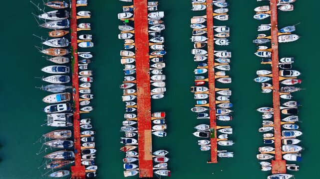 Top view of multiple boat rows docked at marina piers. Aerial perspective of many boats moored at long red piers in turquoise water.