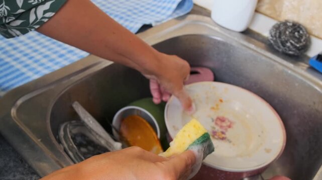 Side view of woman washing dirty dishes in kitchen sink using sponge and detergent. Everyday home routine, dishwashing process, cleaning cookware, domestic chores and hygiene concept.