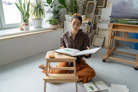 Focused woman artist working on new drawings at home workshop, sitting on floor looking through paper album with watercolor sketches, creative female artisan checking sketchbook with drawing ideas.