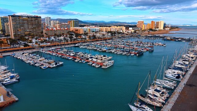 Mediterranean marina with yachts and city skyline background. High angle view of harbor docks filled with boats against a backdrop of mountains and city.
