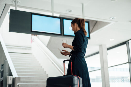 Businesswoman using smart phone and standing near arrival departure board