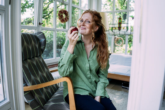 Smiling woman with long hair holding apple and sitting on chair at home