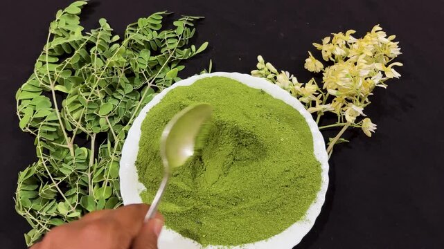 moringa leaf powder in bowl with fresh leaves and flowers on black background