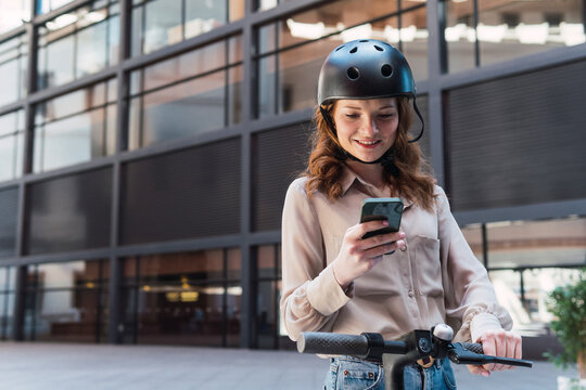 Happy young woman using smartphone and holding electric bicycle in city