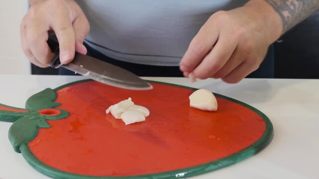 male hands chopping garlic with knife front view on cutting board, mincing cloves into small pieces for cooking, food preparation process, kitchen routine, ingredient ready for recipe use