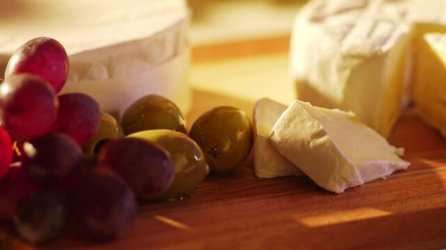 Hand reaching for a green olive on a rustic wooden board with red grapes camembert cheese and mixed olives in warm golden light