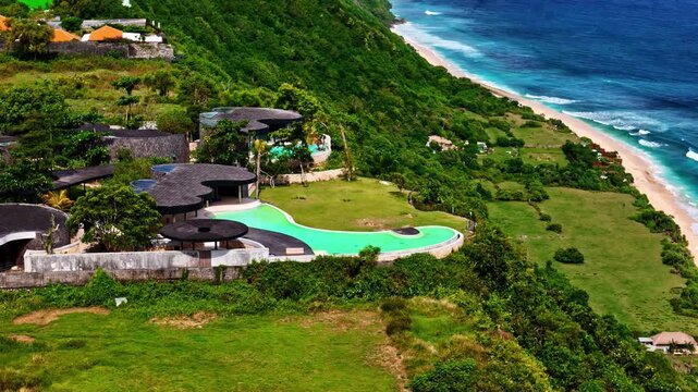 Aerial of an ultra-luxury clifftop villa with winding infinity pool perched above turquoise ocean and white sand beach. Bali, Indonesia, Villa Tebing.