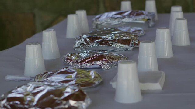 Plastic cups and foil-wrapped plates neatly arranged on a white tablecloth outdoors, creating a ready and welcoming setting for a picnic or event.