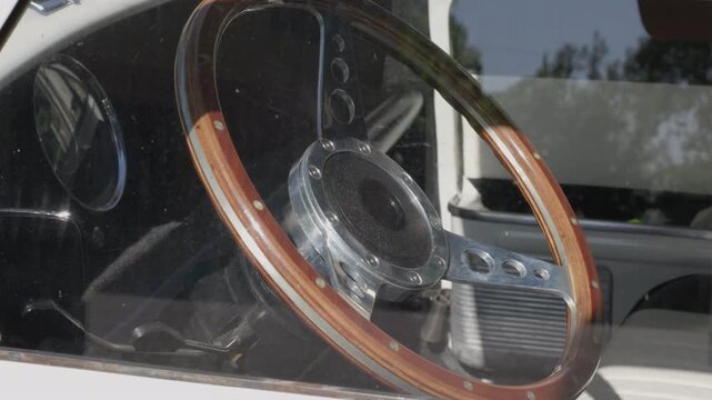 Detailed close-up shot through a window showing a retro wooden steering wheel and dashboard in a sunlit vintage car interior, no people visible.