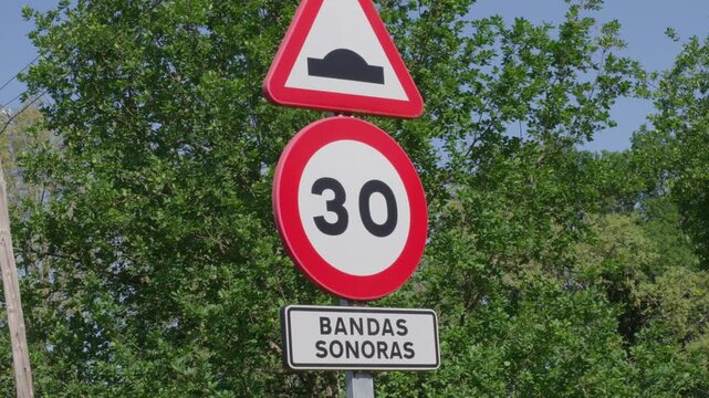 Clear road signs indicate a 30kmh limit and speed bump ahead, bandas sonoras in spanish, under bright daylight, set against leafy green rural scenery.