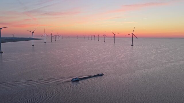 Aerial view of a large cargo barge sailing through an offshore wind farm during a colorful sunset. Calm water reflects the beautiful sky