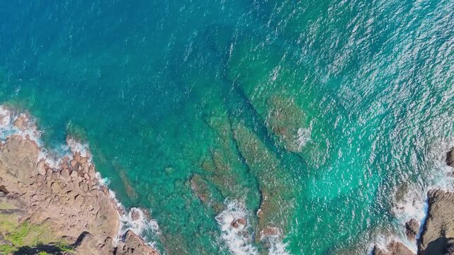 Top-down aerial view of ocean water revealing underwater rock formations along tropical coastline