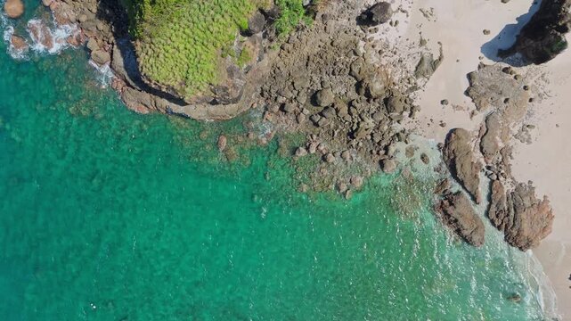 Top-down aerial view of crystal ocean waves crashing against rocky shoreline and sand beach
