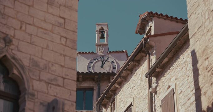 View of a clock tower with a bell and clock face framed by surrounding stone buildings along a narrow street.