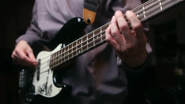 Close up bass player hands plucking and fretting strings of electric bass guitar, rehearsing song or performing live on stage during concert in dark studio. Musician playing electric bass guitar