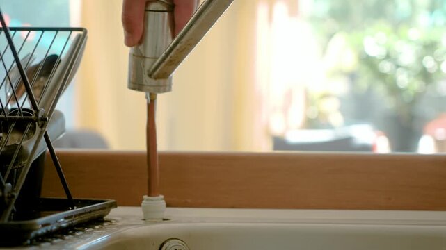 Removing broken kitchen faucet from sink, DIY plumbing repair process at home. Hand unscrewing old tap fixture near drain, maintenance tools and domestic interior background. Fixing water supply