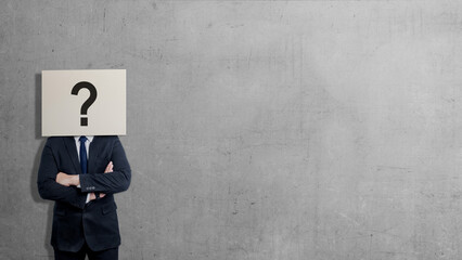 A businessman in a suit stands with arms crossed against a gray concrete wall, his head obscured by a sign featuring a large black question mark, symbolizing anonymity, doubt, or curiosity © Leo Lintang
