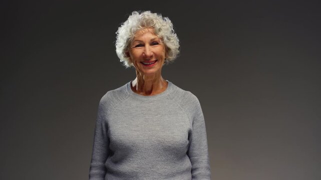 Serious caucasian older retired woman getting happy, positive, cheerful, looking at camera with toothy smile, laughing. Senior female smiles, laughs. Head shot portrait. Happy person in retirement