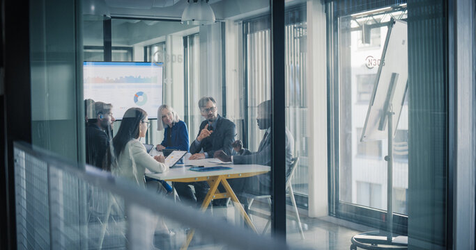 Business Leaders Sit Around a Conference Table in a Glass Meeting Room, Reviewing Charts on a Screen, Discussing Performance, Setting Goals, and Coordinating Strategy for Digital Projects