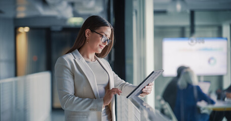 Businesswoman Uses a Digital Tablet in a Modern Office Hallway to Review Project Dashboards, Update Tasks, and Track Performance While a Team Collaborates in a Glass Meeting Room © Gorodenkoff