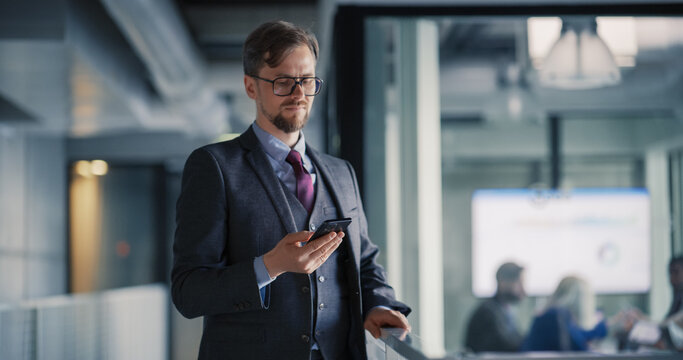 Business Manager Checks a Smartphone Update in a Modern Office Hallway, Reviewing Project Tasks and Live Metrics While a Blurred Team Meets in a Glass Conference Room, Showing Mobile Coordination, Qu