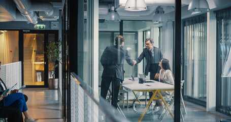 Business Leaders Complete a Job Interview With a Confident Handshake in a Glass Meeting Room, While a Hiring Manager Reviews Notes and Additional Candidates Wait in the Hallway © Gorodenkoff