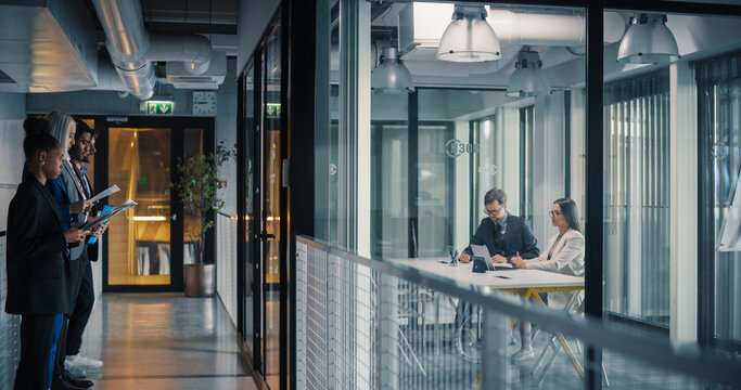 Business Professionals Stand in a Hallway Reviewing Documents and Preparing Pitches, While Two Colleagues in a Glass Meeting Room Analyze Financial Reports on Laptops, Align Priorities