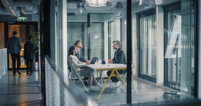 Business Team Sits in a Glass Meeting Room, Discussing Project Plans, Reviewing Reports, and Agreeing on Next Steps While Coworkers Walk Through the Hallway, Creating a Busy Office Atmosphere Focused