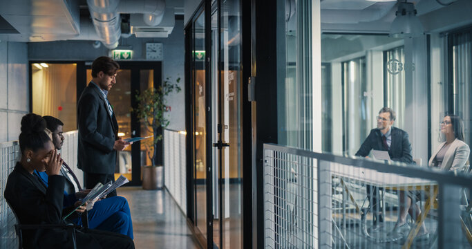 Job Candidates Wait in a Modern Office Corridor Reviewing Resumes and Notes, While Interviewers in a Glass Meeting Room Conduct a Formal Hiring Conversation, Compare Applications, Coordinate Work