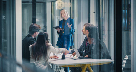 Businesswoman Leads a Strategy Meeting in a Glass Office, Using a Digital Tablet to Explain Goals While a Corporate Team Reviews Data, Negotiates Priorities, Aligns Projects © Gorodenkoff