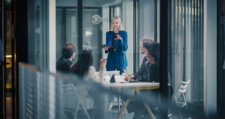 Female Business Leader Stands at the Front of a Glass Conference Room, Using a Digital Tablet to Present Strategy, Guide a Planning Session, Review Performance Metrics, and Coordinate Decisions © Gorodenkoff