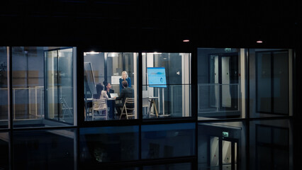 Business Team Works Late in a Glass Office, Reviewing Charts on a Digital Screen, Discussing Project Targets, Adjusting Timelines, and Agreeing Next Steps to Improve Performance, Support Clients © Gorodenkoff