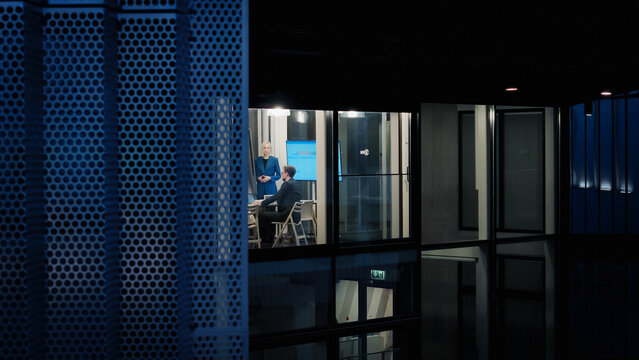Inside a Glass Office at Night, a Female Executive Presents Data on a Large Screen to Seated Colleagues, Explaining Performance Results, Discussing Risks, and Guiding the Team Toward Clear Priorities