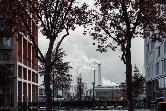 Urban street in Paris with trees and buildings facing an overcast industrial plant where a distant chimney plume marks the city skyline scene