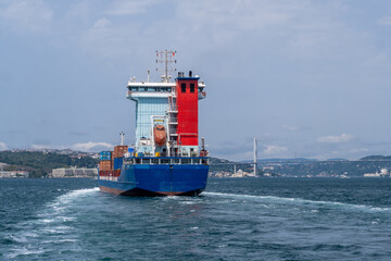 Fototapeta premium A cargo ship moves through the Bosphorus Strait, leaving a wake behind as it approaches the urban scenery, showcasing maritime activity on a sunny day.