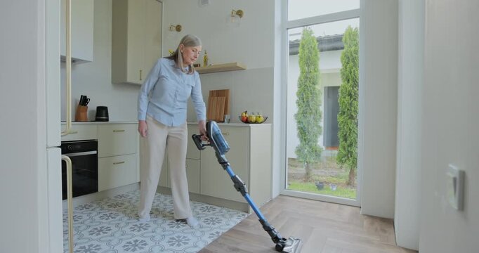Wireless vacuum machine and senior woman cleaning floor in kitchen at home apartment.