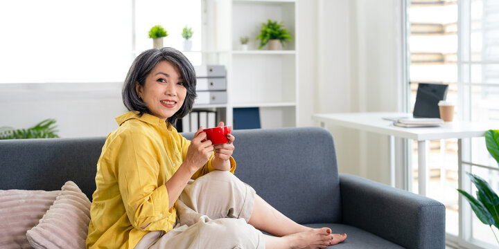 Happy relaxed middle aged asian woman drinking coffee relaxing on sofa at home. Smiling stylish middle aged 50s lady enjoying resting sitting on couch.