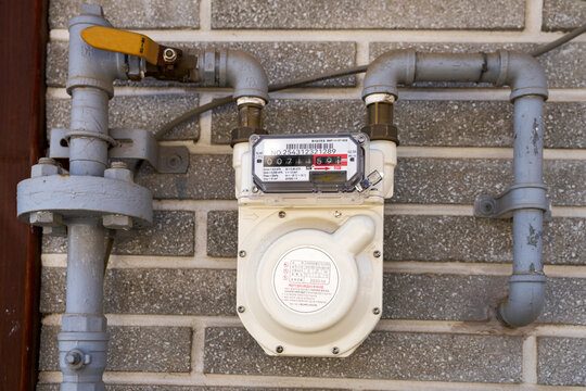Close-up of an outdoor gas utility meter encased in white housing, mounted on brick wall with system of painted metal gas pipe plumbing and valves.